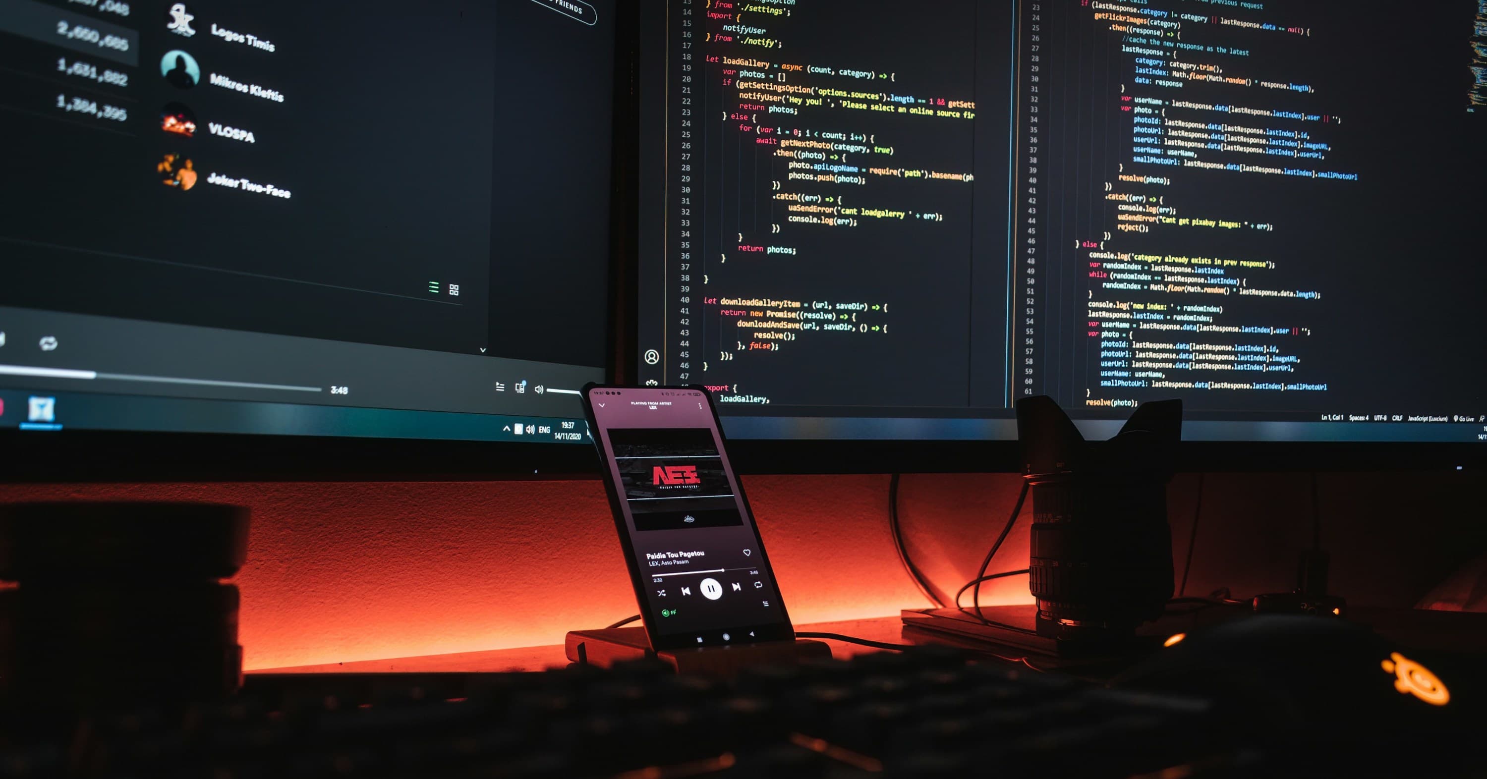 two monitor desk setup with red backlights showing code on one screen and spotify on the other with a phone between them